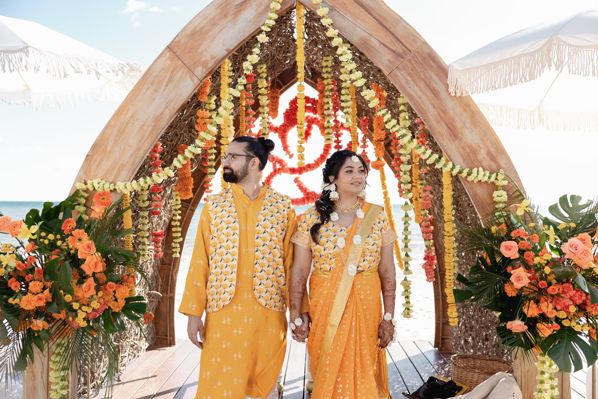 Mihika and Hershel beneath a floral wooden arch by the sea