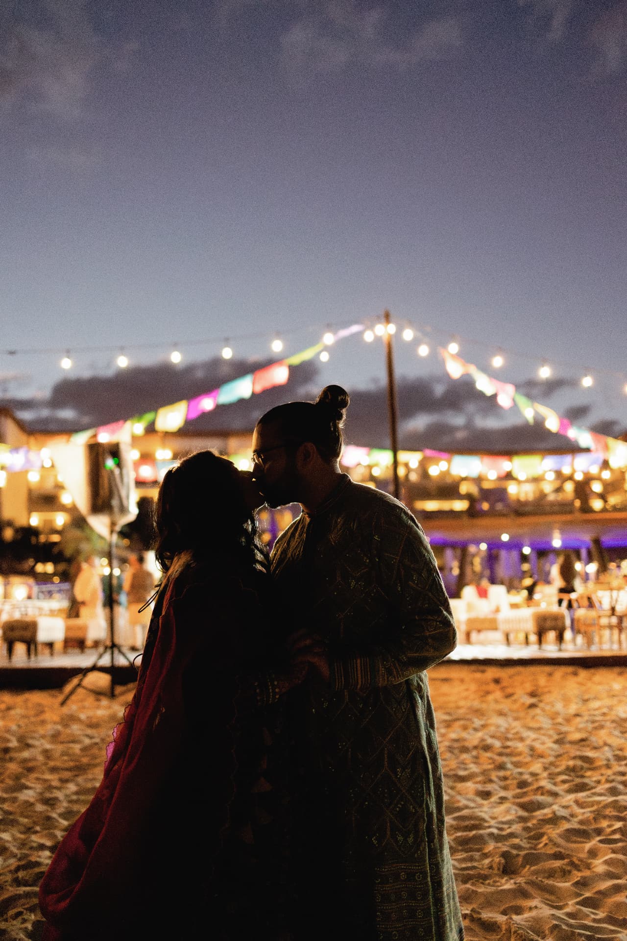The couple silhouetted on the sand beneath string lights and colourful flags