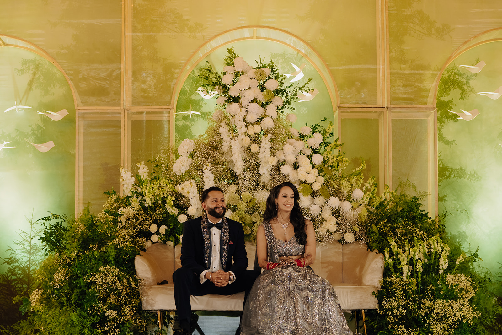 Sneha and Apoorv seated before a white floral installation
